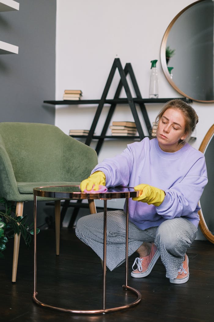 Calm young woman in casual clothes and latex gloves squatting down and wiping table in light living room in daytime